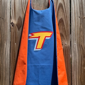 May include: A blue superhero cape featuring a prominent yellow letter T with a red outline and a white shadow effect. The cape has bright orange side panels and a matching orange trim along the edges, hanging vertically against a wooden fence.