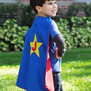 May include: A young boy wearing a blue superhero cape with a red trim and a yellow star with the letter "J" on it. He is wearing a blue shirt and jeans.