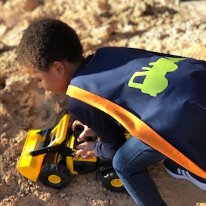 May include: A young child wearing a blue and orange cape with a green tractor design plays with a yellow toy construction vehicle in a sandbox.