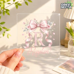May include: A square clear acrylic plaque featuring a large pink letter R and the name Raylan in cursive script. The design includes a prominent pink ribbon bow at the top, surrounded by delicate pink roses and soft green leaves on a transparent base.