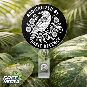 May include: A black and white badge reel featuring the text "RADICALIZED BY BASIC DECENCY" around a raven illustration. The design includes floral details. It has a clear plastic strap and a silver retractable mechanism.