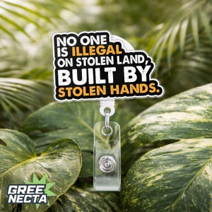 May include: A white and black badge reel with the text "NO ONE IS ILLEGAL ON STOLEN LAND, BUILT BY STOLEN HANDS." The badge reel has a clear plastic holder and a retractable cord. The background is lush green foliage.