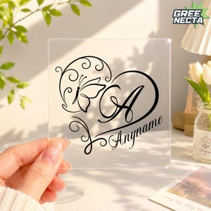 Peut inclure: Une plaque carrée en acrylique transparent présentant un motif noir imprimé d'un papillon et d'une grande lettre A à l'intérieur d'une forme de cœur, avec le texte Anyname écrit en écriture cursive sous la lettre A sur une surface transparente.