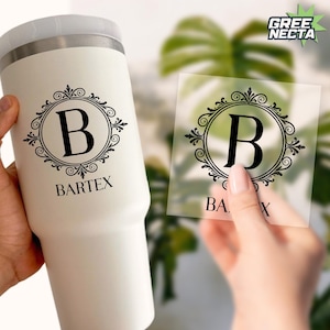 May include: A white insulated tumbler with a black monogram design featuring the letter "B" inside a decorative frame. The word "BARTEX" is printed below the monogram. A clear sticker with the same design is held in a hand.