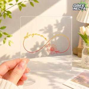 May include: A square clear acrylic plaque featuring a gradient orange to red infinity symbol design. The design includes the text Anyname in a cursive script and a detailed line art daisy flower illustration integrated into the infinity loop.