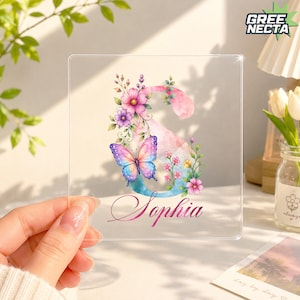 May include: A clear square acrylic plaque featuring a large letter S decorated with watercolor-style pink and purple flowers and a vibrant butterfly. The name Sophia is printed in a cursive script font below the letter in a gradient pink color.