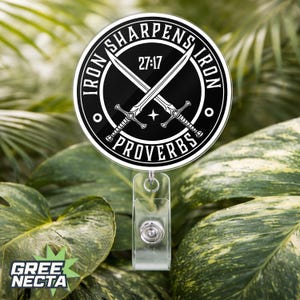 Peut inclure: Un enrouleur de badge rond, noir et blanc, avec l'inscription "IRON SHARPENS IRON" et "PROVERBES 27:17" autour de deux épées croisées. Il est doté d'une lanière en plastique transparent et d'un clip rétractable argenté. Le fond est un feuillage vert flou.