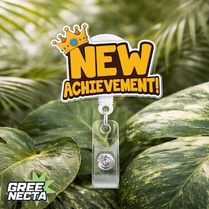 May include: A badge reel with the words "NEW ACHIEVEMENT!" in yellow and brown, topped with a gold crown. The reel has a clear plastic ID holder and a silver metal clip. The background is lush green foliage.