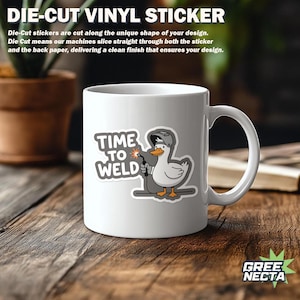 Pu&ograve; includere: Tazza in ceramica bianca con un adesivo di un'anatra saldatore dei cartoni animati con la scritta "TIME TO WELD". L'anatra indossa un casco da saldatore e tiene in mano una torcia. La tazza &egrave; su una superficie di legno.