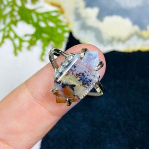 Puede incluir: Un anillo plateado con una gema grande de forma irregular, con colores como marrón, azul y blanco. El anillo tiene un diseño único con pequeñas piedras transparentes.