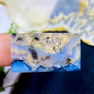 May include: A rectangular gemstone with a landscape pattern. The stone displays light blue, white, beige, and black hues. The design evokes a cloudy sky over water, with tree-like formations. The stone is likely a decorative item.