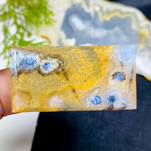Könnte beinhalten: Ein rechteckiger Edelstein mit polierter Oberfläche. Der Stein weist eine Mischung aus Farben auf, darunter Gelb, Weiß, Blau und Braun. Das Muster umfasst kleine, blütenartige blaue Einschlüsse und eine gesprenkelte Textur.