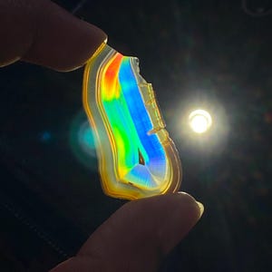 May include: A translucent, irregularly shaped slice of agate held against a bright light source. The stone displays vibrant, glowing bands of yellow, orange, green, and blue, with a distinct rainbow-like spectrum effect visible through the center.