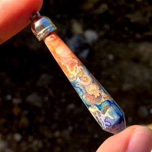 May include: A tapered, cylindrical pendant featuring a multi-colored stone with swirling patterns of orange, cream, blue, and purple. The stone is capped with a polished silver-toned metal bail at the top, designed for attachment to a necklace chain.