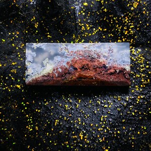 Op de afbeelding: Een rechthoekige, gepolijste steen met een landschapachtig patroon. De steen heeft een mix van kleuren, waaronder tinten bruin, rood, wit en grijs, die lijken op een miniatuur bergketen of wolkenlandschap. De steen is geplaatst tegen een donkere, glinsterende achtergrond.