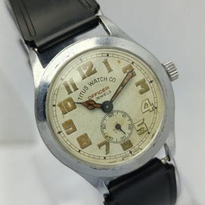 May include: Vintage Titus Watch Co. Officer watch with a silver-tone case and a black leather band. The watch face is off-white with large, brown numerals and hands. The watch has a small seconds dial and the words "TITUS WATCH CO OFFICER JEWELS SWISS MADE" are visible.