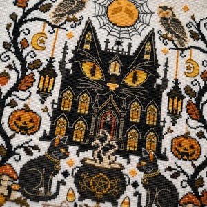 K&ouml;nnte beinhalten: Ein Kreuzstichmuster mit einem schwarzen, katzenf&ouml;rmigen Spukhaus mit leuchtend gelben Augen. Das Design umfasst K&uuml;rbisse, Eulen, einen Kessel und einen Vollmond, alles im Halloween-Thema. Die Farbpalette umfasst Schwarz, Orange und Creme.