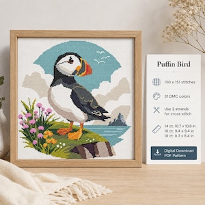 Puede incluir: Un patrón de punto de cruz con un frailecillo en un marco de madera. El diseño muestra un frailecillo en un acantilado con flores rosas y amarillas, cielo azul y nubes. El texto incluye Puffin Bird, 150 x 151 puntadas, 31 colores DMC y dimensiones en cm para telas de 14, 16 y 18 ct.