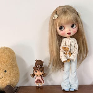 May include: A doll with long blonde hair, wearing a white floral long-sleeve top and light blue trousers, holding a small brown teddy bear. Beside it stands a smaller doll wearing a red and white striped shirt with a brown bear figure on its head.