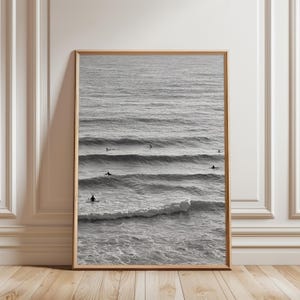 May include: A framed monochrome photograph of the sea with surfers riding waves. The image is in a wooden frame, leaning against a white wall and a wooden floor.