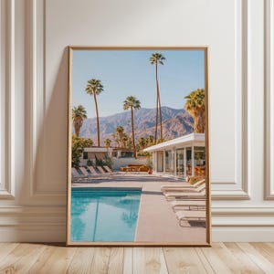 May include: A framed print showing a poolside scene with palm trees and mountains under a blue sky. The pool reflects the sky, and sun loungers are arranged nearby. A white building with large windows is also visible in the distance.