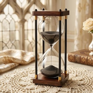 May include: An hourglass with a dark wood frame and clear glass bulbs filled with black sand. The hourglass has gold accents and is set on a wooden base. The background includes a lace doily and a decorative window.