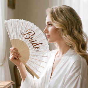 May include: A handheld folding fan featuring white lace fabric and a light wood frame, with the word Bride written in a dark brown cursive script across the center of the lace. The fan is held upright, displaying the intricate lace pattern.