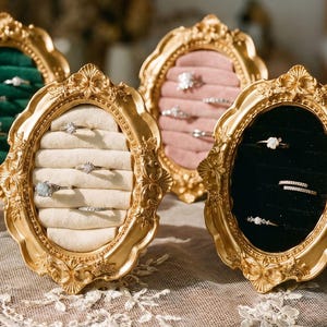 Könnte beinhalten: Verzierte goldgerahmte Schmuckdisplays mit Samtinnenräumen in Rot, Grün, Rosa, Creme und Schwarz. Die Displays halten Ohrringe und Ringe und präsentieren verschiedene Schmuckstile. Die Rahmen stehen auf einer spitzenbedeckten Oberfläche.