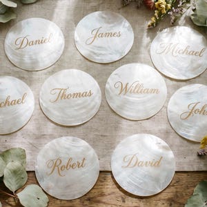 Könnte beinhalten: Neun runde, weiße Tischkarten mit goldenen Namen: Daniel, James, Michael, Thomas, William, John, Robert und David. Sie liegen auf einem beigen Tischläufer, umgeben von floralen und grünen Dekorationen.