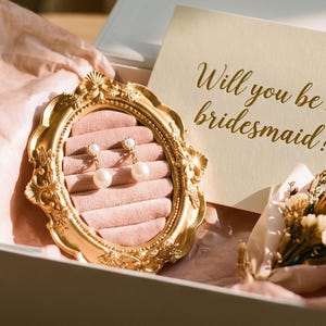 Könnte beinhalten: Geschenkbox mit einem goldgerahmten Schmuckhalter mit Perlenohrringen. Eine Karte mit dem Text "Will you be my bridesmaid?" und ein kleiner Strauß Trockenblumen befinden sich ebenfalls in der Box. Die Box ist mit rosa Seidenpapier ausgekleidet.