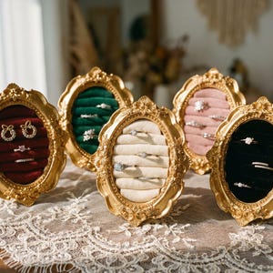Könnte beinhalten: Verzierte goldgerahmte Schmuckdisplays mit Samtinnenräumen in Rot, Grün, Rosa, Creme und Schwarz. Die Displays halten Ohrringe und Ringe und präsentieren verschiedene Schmuckstile. Die Rahmen stehen auf einer spitzenbedeckten Oberfläche.