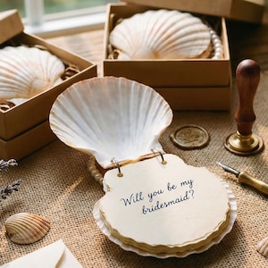 Peut inclure: Une invitation en forme de coquillage avec le texte "Will you be my bridesmaid?" est ouverte sur une table en bois. Deux coquillages sont dans de petites boîtes marron. Un bouquet de lavande, un stylo et un sceau de cire sont également sur la table.