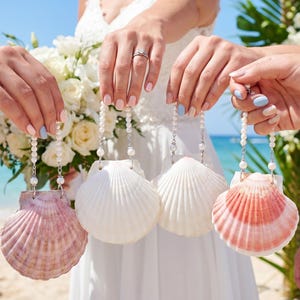 Puede incluir: Cuatro monederos de conchas marinas en varios colores rosa y blanco son sostenidos por personas. Cada monedero está suspendido de una cadena de cuentas. Un ramo de flores blancas se sostiene en el fondo. La escena tiene lugar en una playa.