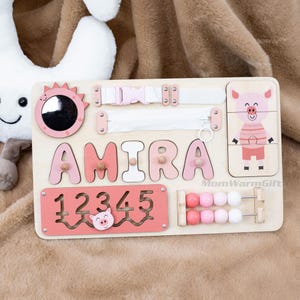 May include: Wooden activity board with the name "AMIRA" in pink letters, a mirror, a zipper, a buckle, a pig puzzle, and an abacus. The board also features a pink rectangle with the numbers 1 through 5 and a pig graphic. The text "MomWarmGift" is also visible.