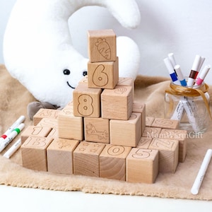 May include: Wooden alphabet blocks with numbers and illustrations, including a carrot, giraffe, and stars. The blocks are arranged on a soft, brown surface, with markers and a plush moon in the background. The blocks are a natural wood color.