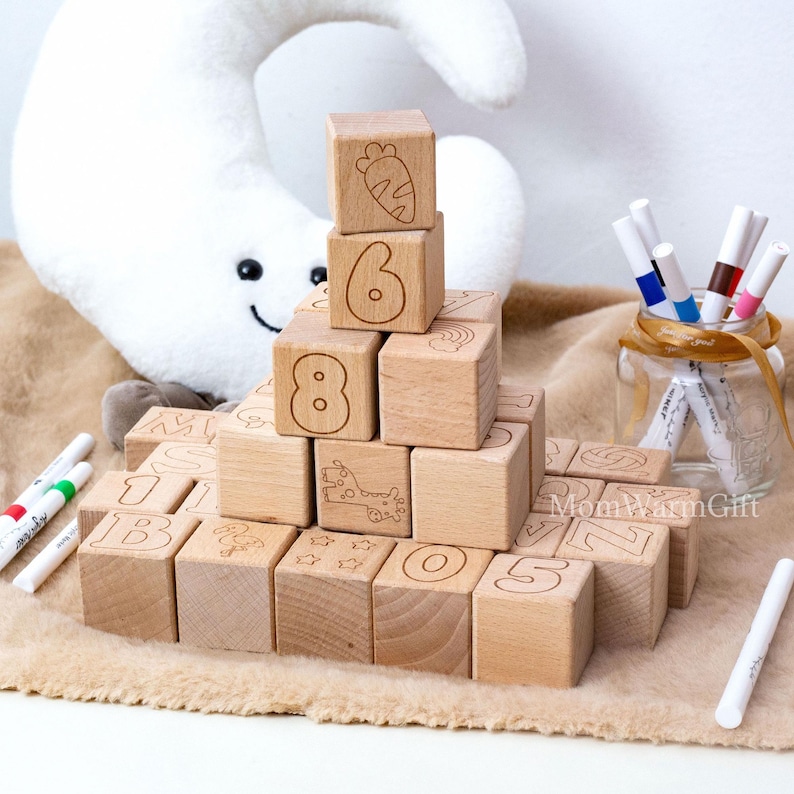 May include: Wooden alphabet blocks with numbers and illustrations, including a carrot, giraffe, and stars. The blocks are arranged on a soft, brown surface, with markers and a plush moon in the background. The blocks are a natural wood color.