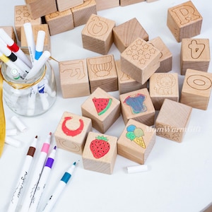 May include: Wooden alphabet blocks with engraved letters, numbers, and images of a lion, watermelon, and ice cream. The blocks are accompanied by a set of colorful acrylic markers in a glass jar.