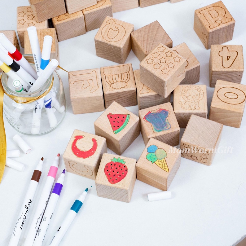 May include: Wooden alphabet blocks with engraved letters, numbers, and images of a lion, watermelon, and ice cream. The blocks are accompanied by a set of colorful acrylic markers in a glass jar.