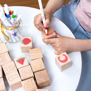 May include: Wooden alphabet blocks with painted letters and fruit illustrations. A child is using a red marker to draw on a block. A jar of colorful markers sits nearby. The blocks are on a white table.