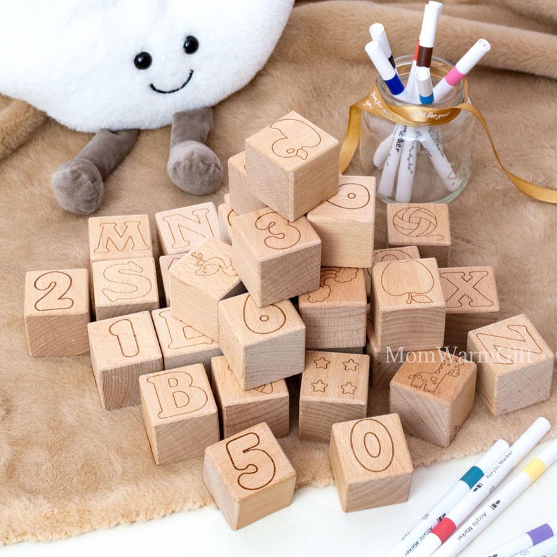 May include: Wooden alphabet and number blocks with engraved letters, numbers, and simple illustrations. A plush cloud toy and a glass jar with markers are in the background. The blocks are scattered on a soft, tan surface.