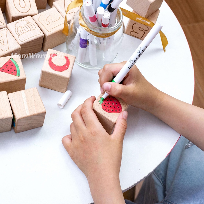 May include: Wooden alphabet blocks with painted illustrations, including a red strawberry, a watermelon slice, and a crescent moon. A hand is using a white marker to add details to a strawberry illustration on a block. A jar of markers sits nearby.