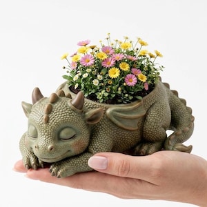 May include: A green dragon-shaped planter with a textured surface, holding a vibrant arrangement of pink and yellow flowers. The dragon is in a sleeping pose, with closed eyes and small horns. The planter is held in a hand.