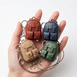 May include: Four miniature backpack keyrings in red-brown, navy blue, tan, and green. Each has two front pockets with heart-shaped details and silver-tone hardware. The keyrings are held in a hand against a white background.
