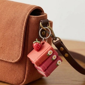 May include: A brown canvas bag with a leather strap and a keychain. The keychain features a red strawberry charm and a stack of miniature books in shades of pink, adorned with small white flowers.