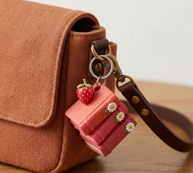 May include: A brown canvas bag with a leather strap and a keychain. The keychain features a red strawberry charm and a stack of miniature books in shades of pink, adorned with small white flowers.