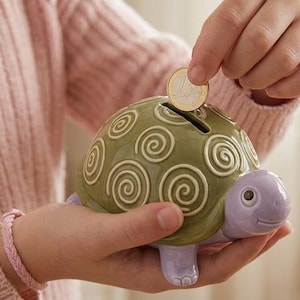 May include: A ceramic turtle-shaped piggy bank in shades of green and purple. The turtle's shell features a spiral design. A coin is being inserted into the slot on top. The piggy bank is held in someone's hands.
