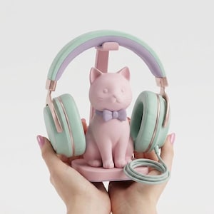 May include: Mint green and lilac headphones with rose gold detailing sit on a pink cat-shaped stand. The cat has a purple bow tie. The headphones are wired.