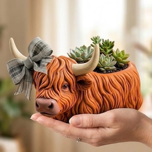 Può includere: Una fioriera in ceramica a forma di mucca delle Highlands nei toni dell'arancione e del marrone, ornata da un fiocco a quadri. La fioriera è riempita di piccole piante grasse verdi. La mucca ha grandi corna.