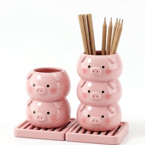 May include: Two pink ceramic containers shaped like stacked pig heads, one with two tiers and one with three tiers, resting on matching pink slatted rectangular bases. The taller container holds several light brown wooden pencils.