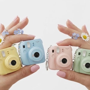 May include: Four miniature pastel cameras in yellow, light blue, pink, and mint green. Each camera has a black lens and a silver keychain. The cameras are held up by hands decorated with flowers.